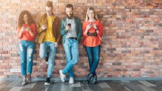 Millennials social media addicted generation. Young people in colorful outfits leaned against brick wall, using smartphones.