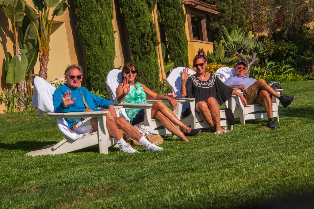 Group of two couples of baby boomers relaxing and waving while sitting on white adirondack on luxurious green grass lawn