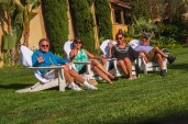 Group of two couples of baby boomers relaxing and waving while sitting on white adirondack on luxurious green grass lawn