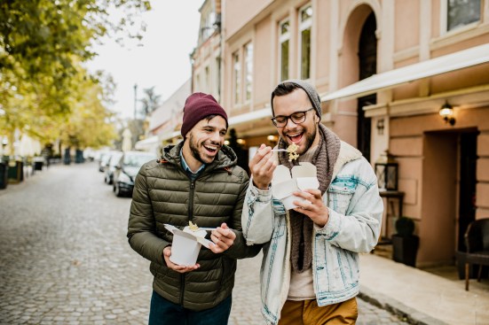 Two male friends are outdoors, eating pasta to go and talk